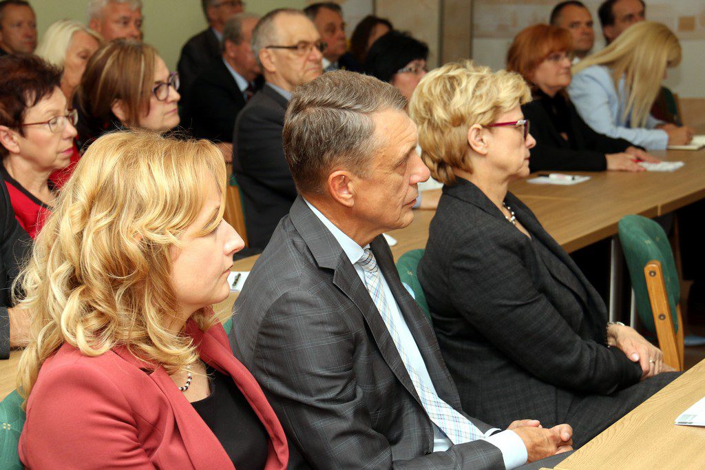 Ludzie siedzą przy stołach, słuchając. Widać kobiety i mężczyzn w formalnych strojach.