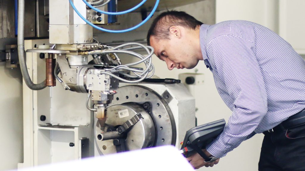 Mężczyzna w kratę obsługuje maszynę CNC. Kluczowe obiekty: mężczyzna, maszyna CNC, panel sterowania.