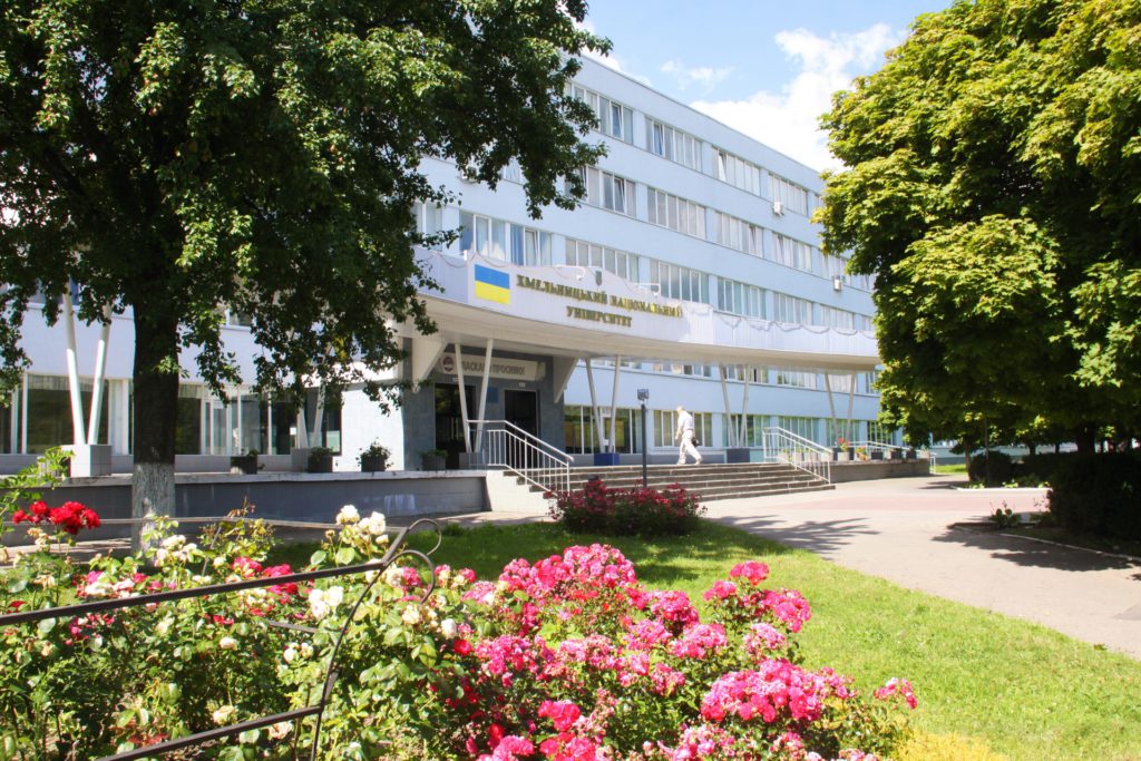 Budynek uniwersytetu z flagą Ukrainy. Kluczowe obiekty: budynek, drzewa, kwiaty, schody, osoba. Kontekst: teren uczelni.