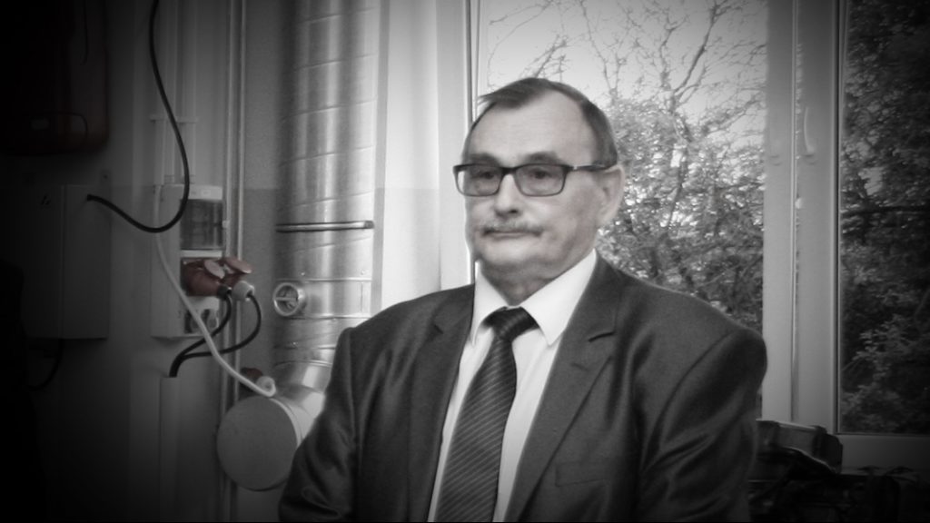 Mężczyzna w okularach i garniturze stoi obok okna. W tle widoczne są rury i drzewa.