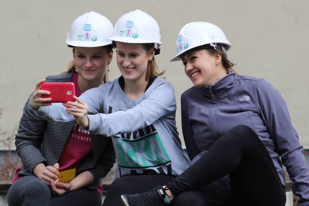 Trzy kobiety w białych kaskach robią selfie. Kobiety, kaski, telefon, kurtki.