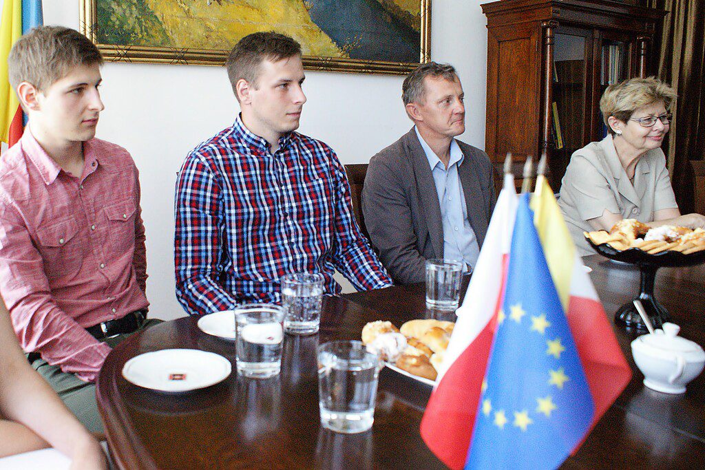 Cztery osoby siedzą przy stole. Na stole są flagi Polski i UE, ciastka, talerzyki i szklanki z wodą.