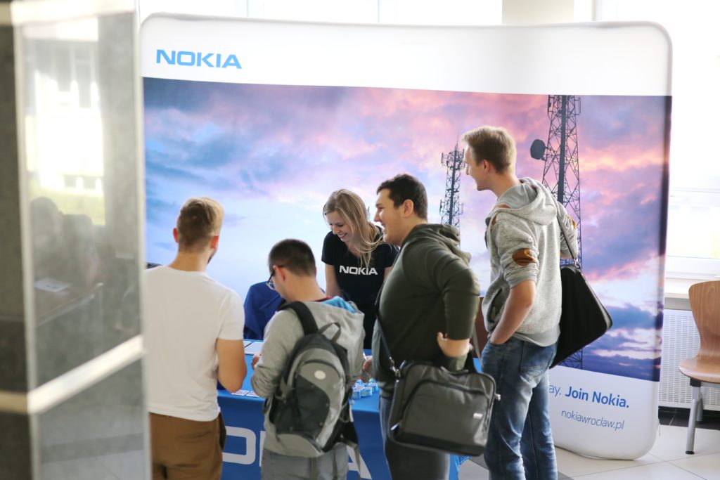 Ludzie przy stoisku Nokia. Tło z logiem Nokia i zdjęciem masztów telekomunikacyjnych.