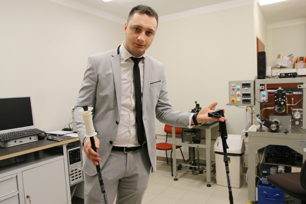 Mężczyzna w garniturze trzyma kijki trekkingowe. W tle widać sprzęt laboratoryjny i komputer.