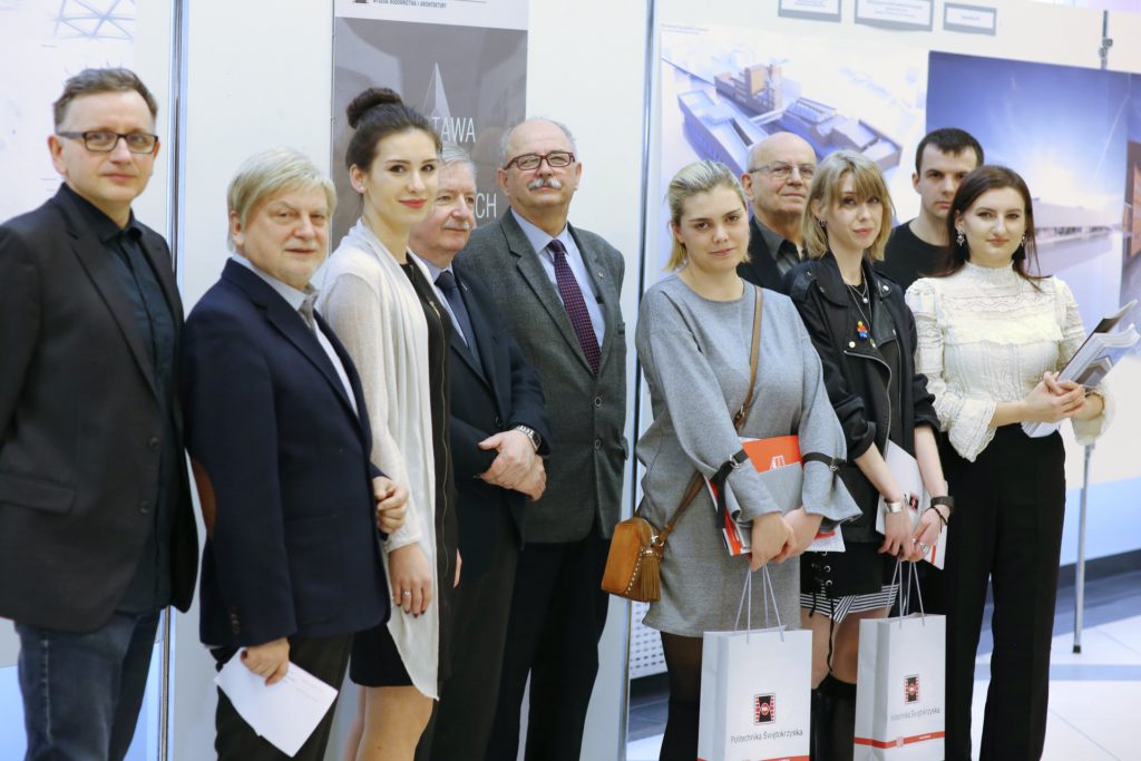 Grupa osób pozuje do zdjęcia. Widać mężczyzn i kobiety w eleganckich strojach, stojących przed tablicami informacyjnymi.