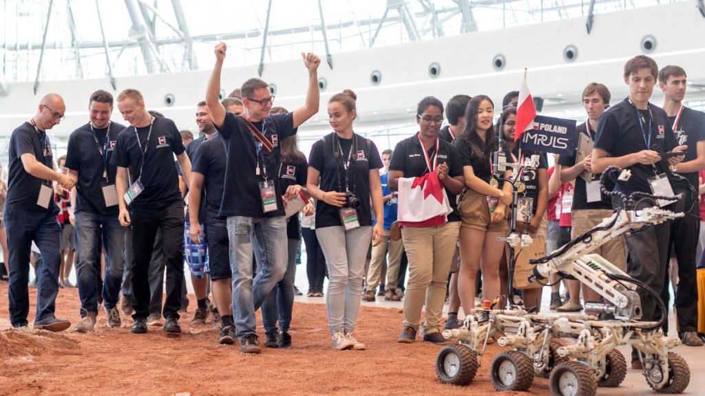 Grupa młodych ludzi w czarnych koszulkach z identyfikatorami, z flagą Polski i robotem. Konkurs robotyki.