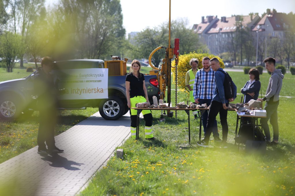 Grupa osób przy samochodzie z logo Politechniki Świętokrzyskiej i stołach z próbkami skał.