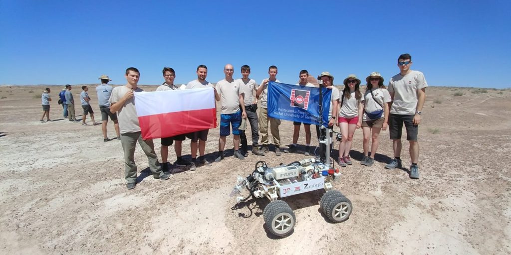 Grupa ludzi stoi na pustyni z polską flagą i robotem. Kluczowe obiekty: ludzie, robot, flaga Polski, pustynia.
