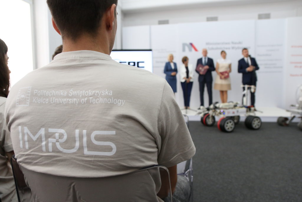 Osoba w koszulce z logo Politechniki Świętokrzyskiej. W tle panel z logo, osoby i robot.