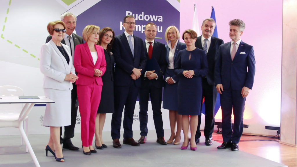 Grupa osób w eleganckich strojach stoi przed banerem z napisem 