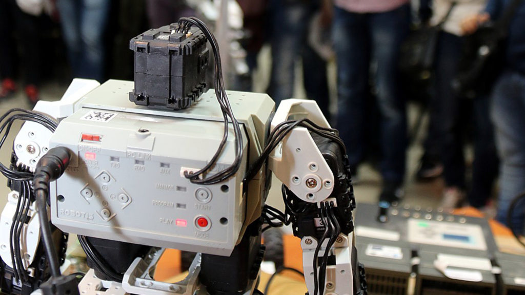 Robot z modułem sterującym i okablowaniem. W tle widać ludzi.