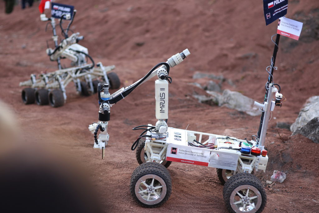 Dwa roboty terenowe z ramionami i flagami Polski. Jeden z nich ma napis 
