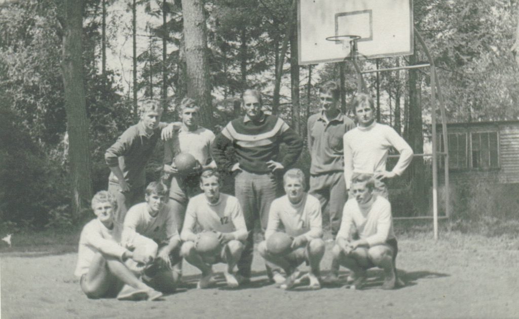 Stare zdjęcie. Grupa młodych mężczyzn pozuje do zdjęcia na boisku sportowym. W tle widać drzewa i budynek.