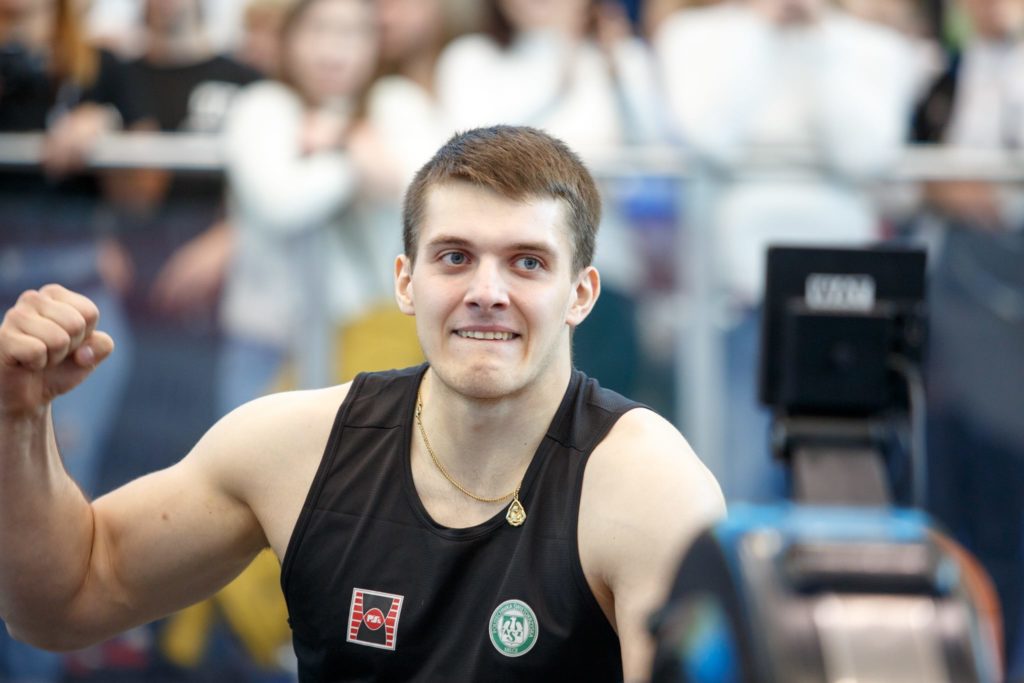 Młody mężczyzna w czarnej koszulce sportowej z emblematami, zaciśnięta pięść. Kontekst: zawody sportowe.