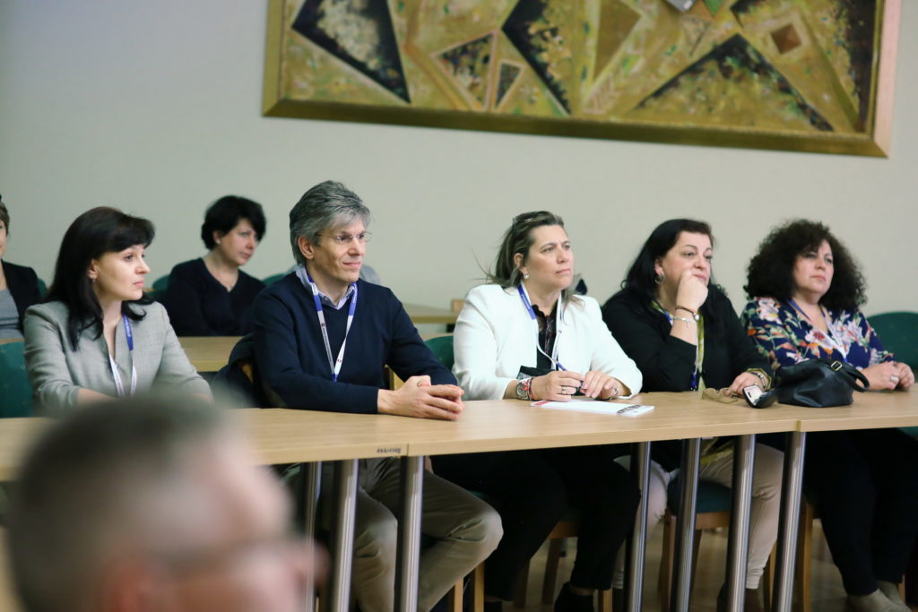Ludzie siedzą przy stołach podczas konferencji. Kluczowe obiekty: osoby, stoły, krzesła, obraz na ścianie.