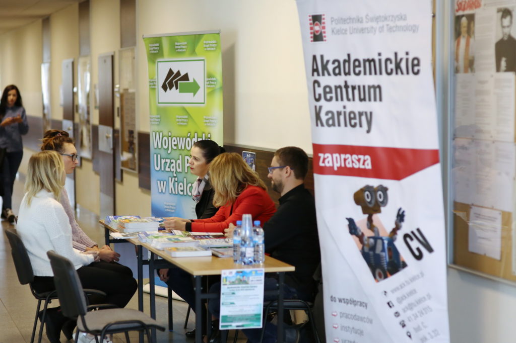 Ludzie siedzą przy stołach, obok banerów informacyjnych. Kontekst: targi pracy lub wydarzenie edukacyjne.