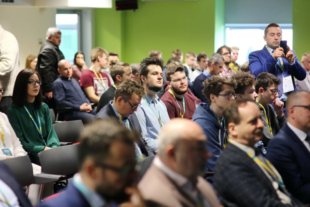 Ludzie siedzą na sali konferencyjnej. Widać mężczyznę robiącego zdjęcie telefonem.