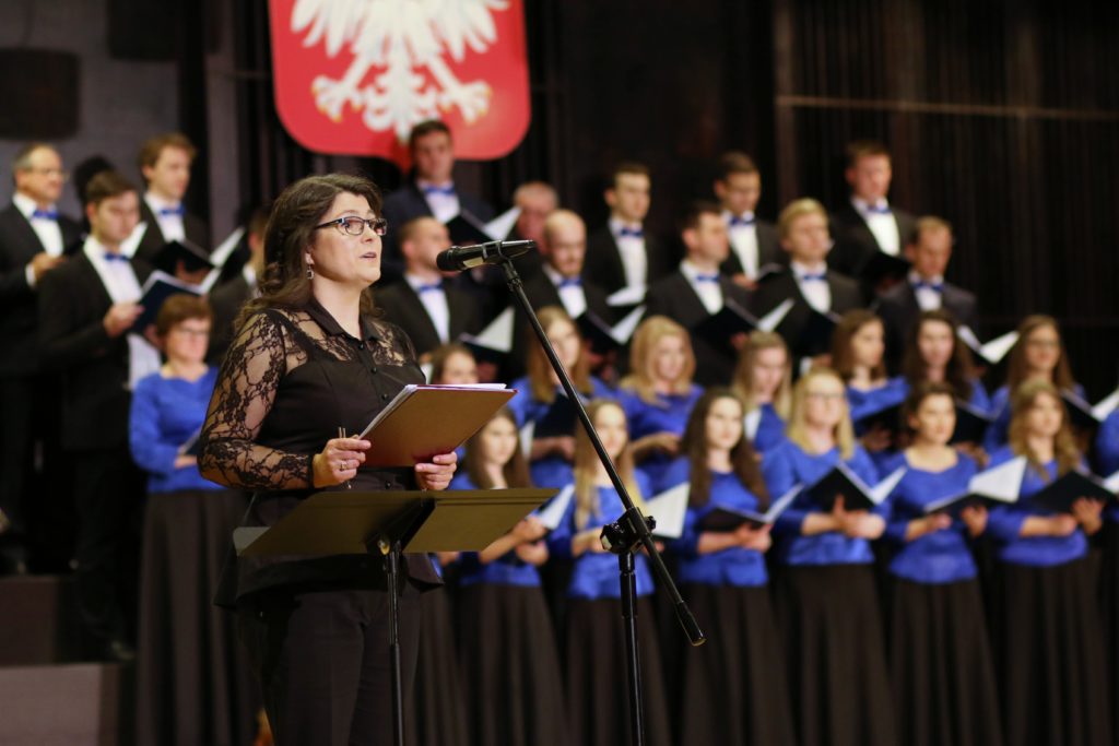 Kobieta z nutami śpiewa przed chórem. W tle godło Polski.