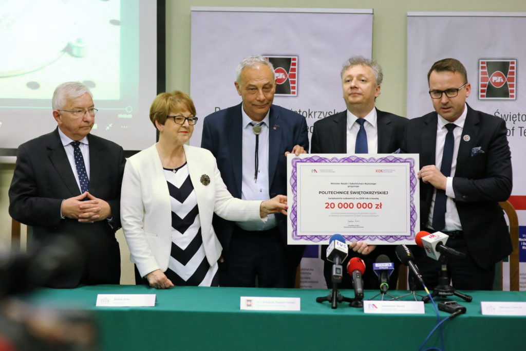 Cztery osoby stoją za zielonym stołem, trzymając certyfikat. Kluczowe obiekty: certyfikat, mikrofony, ludzie.