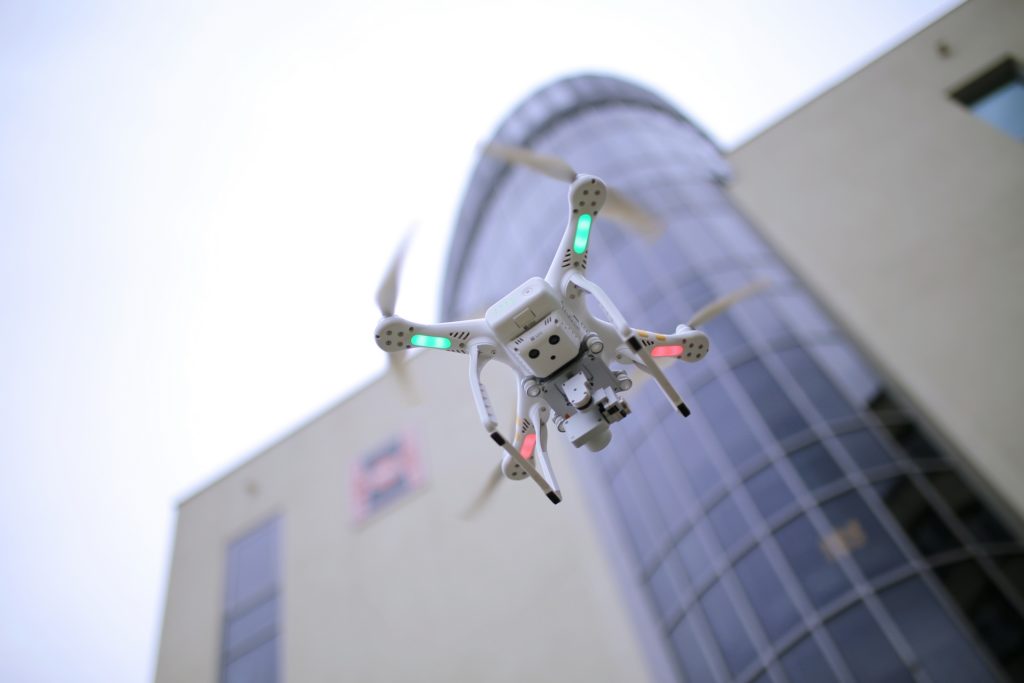Biały dron z zielonymi i czerwonymi światłami leci przed nowoczesnym budynkiem.