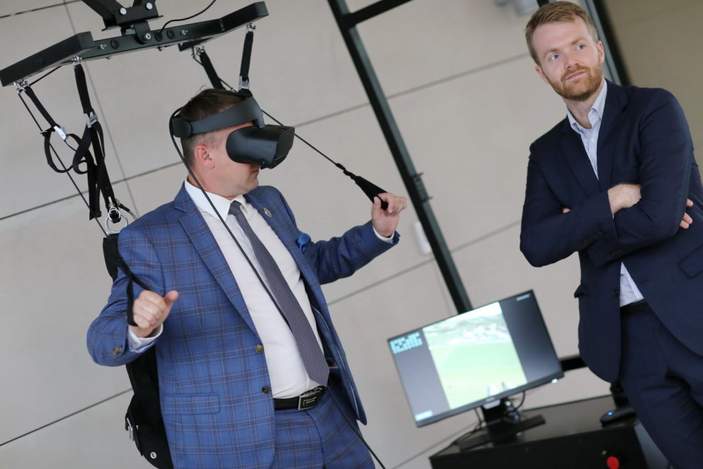 Mężczyzna w goglach VR i garniturze testuje symulator. Obok stoi drugi mężczyzna w garniturze.