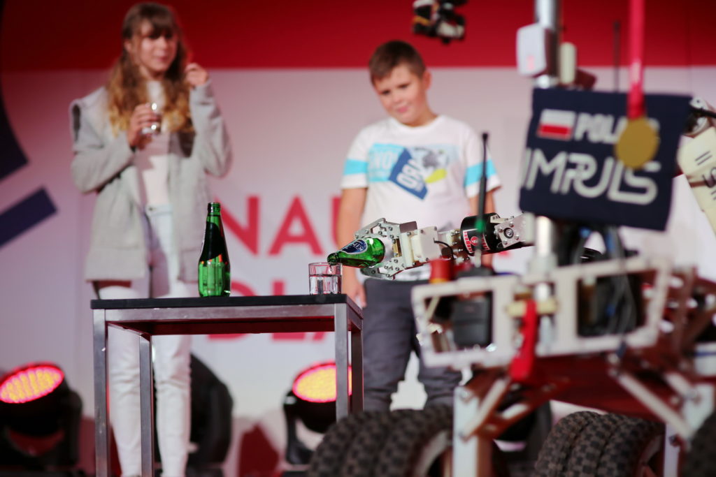 Robot nalewa płyn z butelki do szklanki. W tle dziewczyna i chłopiec.