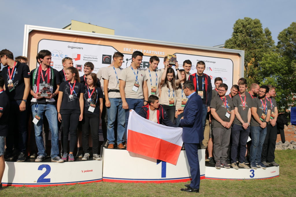 Grupa młodych ludzi na podium z pucharem i flagą Polski. Kontekst: ceremonia wręczenia nagród.