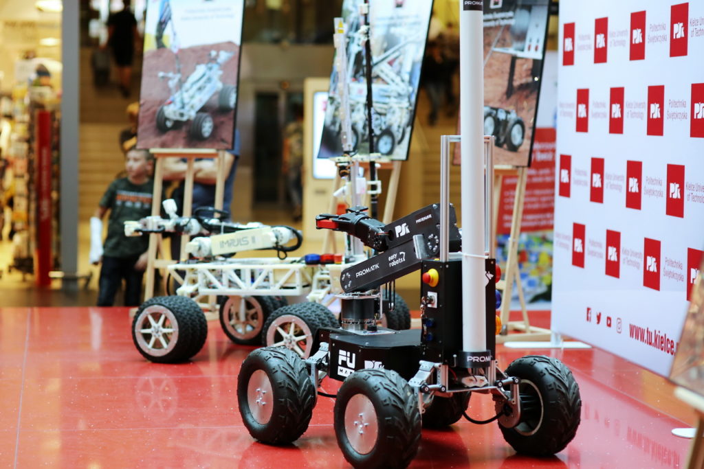 Dwa roboty mobilne z ramionami, jeden biały, drugi czarny, na czerwonej podłodze. W tle wystawa z plakatami.