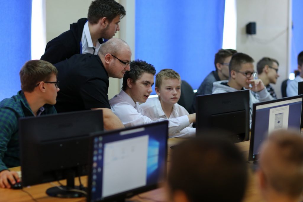 Grupa osób, w tym młodzież i dorośli, pracuje przy komputerach w sali lekcyjnej.