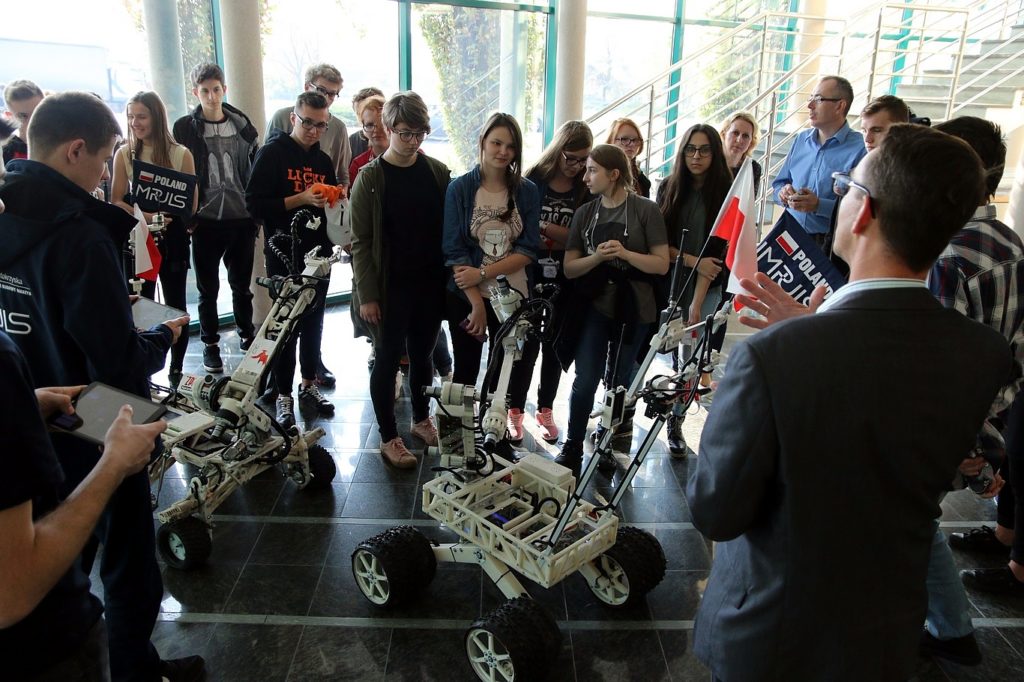 Grupa młodych ludzi ogląda roboty. W tle widać flagę Polski.