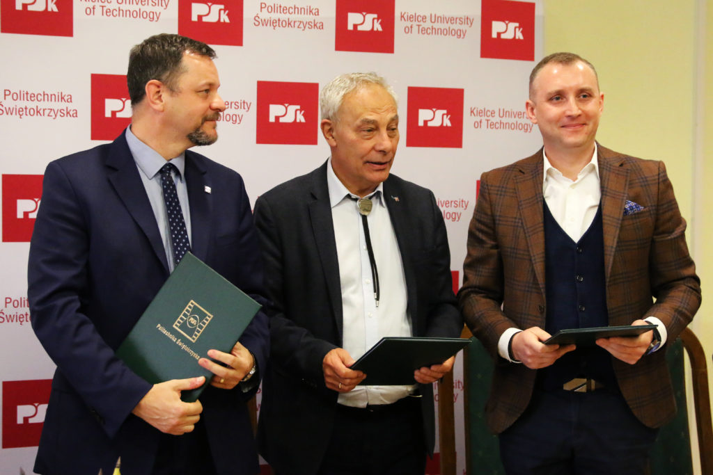 Trzech mężczyzn w garniturach stoi przed ścianą z logo Politechniki Świętokrzyskiej. Trzymają zielone teczki.