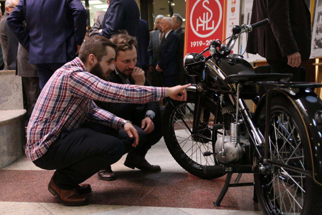 Dwóch mężczyzn ogląda zabytkowy motocykl. W tle widoczny baner z datą 1919-2019.