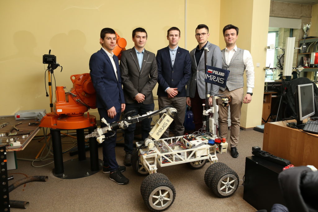 Pięciu mężczyzn stoi obok robota mobilnego i ramienia robota. Kontekst to prawdopodobnie projekt inżynieryjny lub edukacyjny.