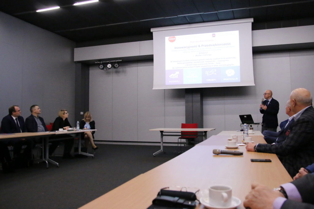 Konferencja z prezentacją na ekranie. Uczestnicy siedzą przy stołach, jeden mężczyzna przemawia.