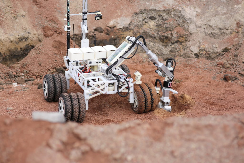 Robot z ramieniem i kamerą na kołach, pracujący na czerwonej, piaszczystej powierzchni.