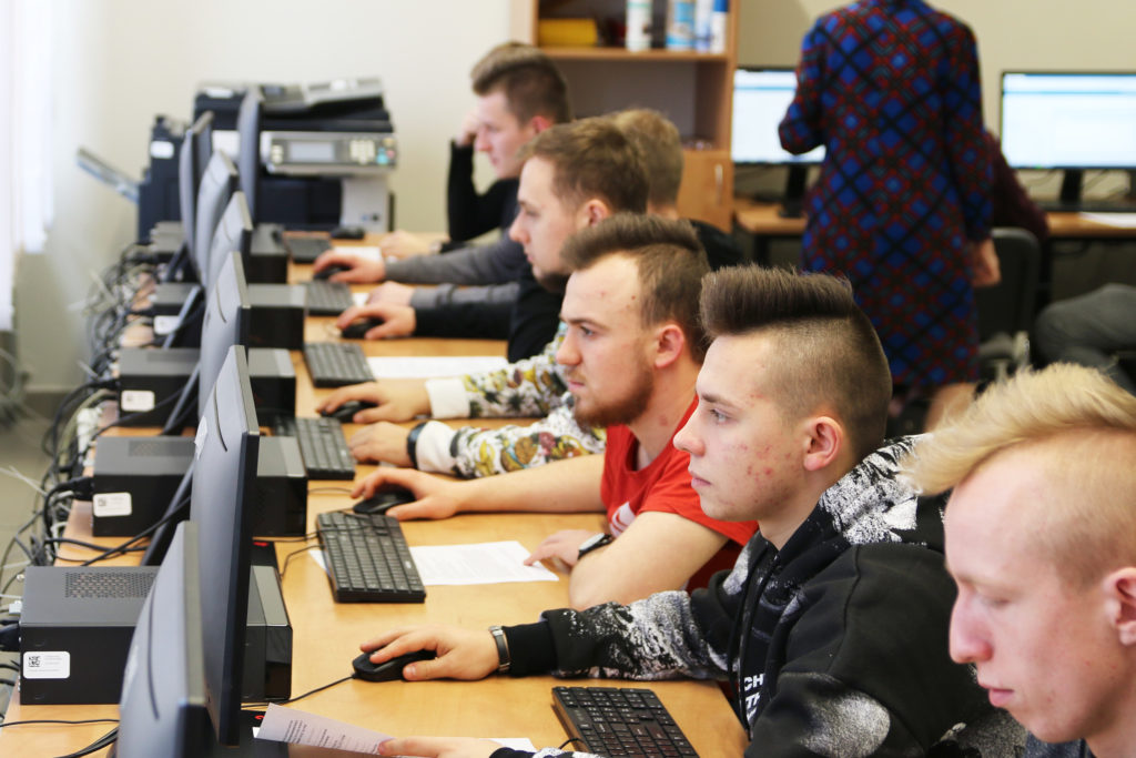 Uczniowie siedzą przy komputerach w sali lekcyjnej. Kluczowe obiekty: monitory, klawiatury, myszki, uczniowie.