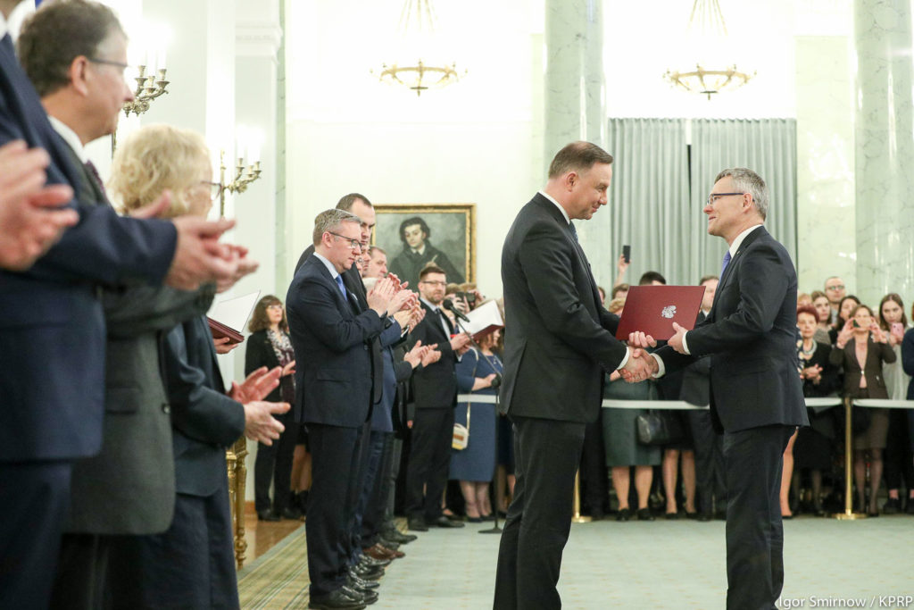 Prezydent RP wręcza dokument. W tle stoją ludzie, w tym mężczyzna z dyplomem.