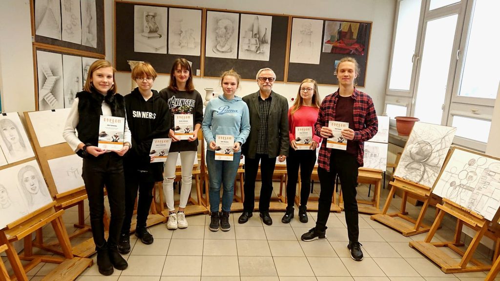 Grupa osób z dyplomami stoi przed pracami plastycznymi. W tle rysunki i szkice.