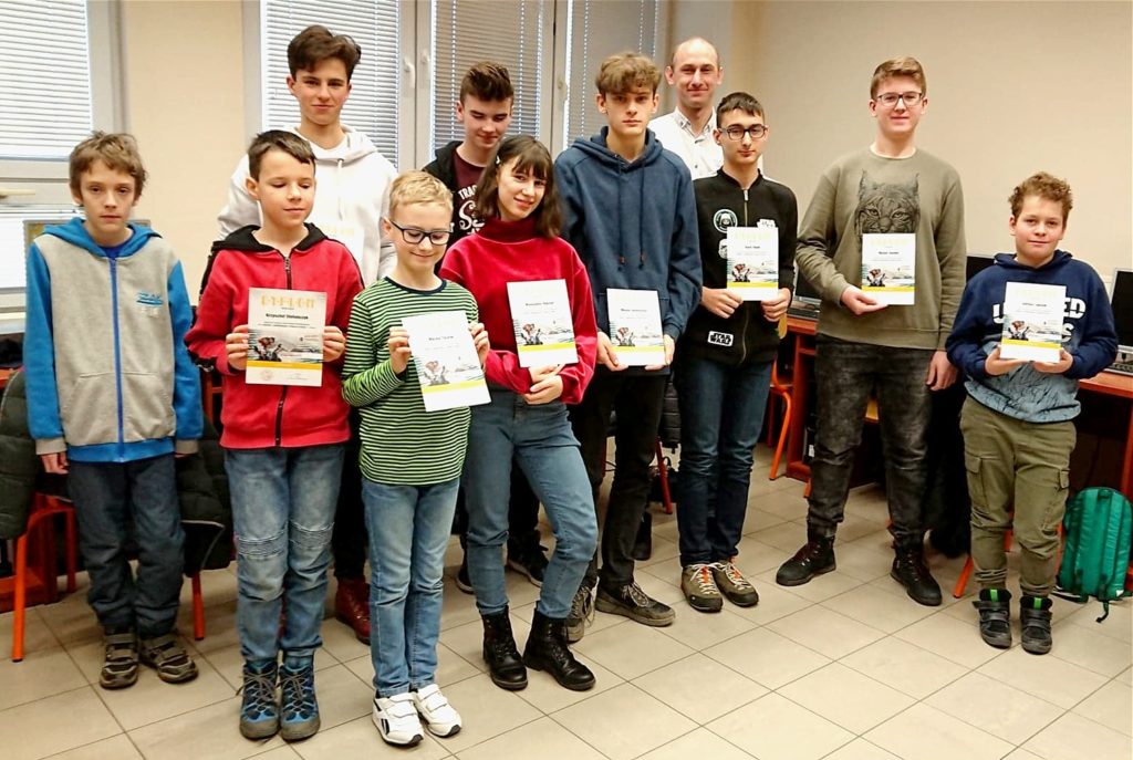 Grupa dzieci i dorosłych pozuje do zdjęcia, trzymając certyfikaty. Wszyscy są w pomieszczeniu.