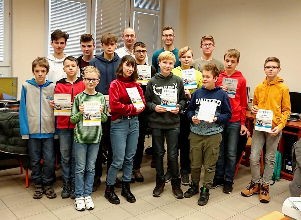 Grupa młodzieży i dorosłych pozuje do zdjęcia, trzymając certyfikaty. Kluczowe obiekty: ludzie, certyfikaty, komputery.