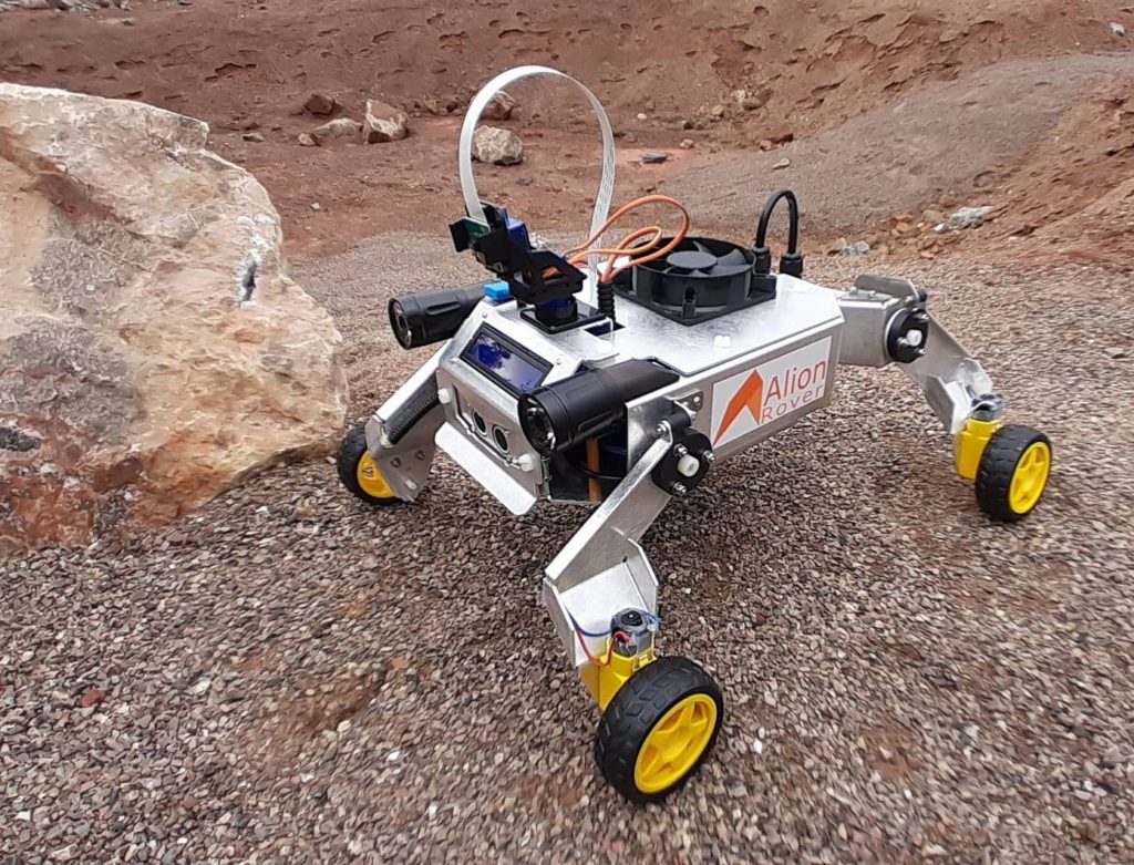 Sześciokołowy łazik "Alion Rover" z kamerami i wentylatorem na kamienistym, czerwonym podłożu.