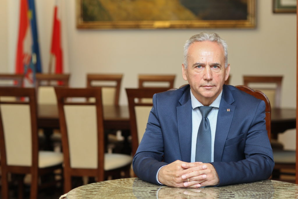 Mężczyzna w garniturze siedzi przy stole. W tle flagi i obrazy.