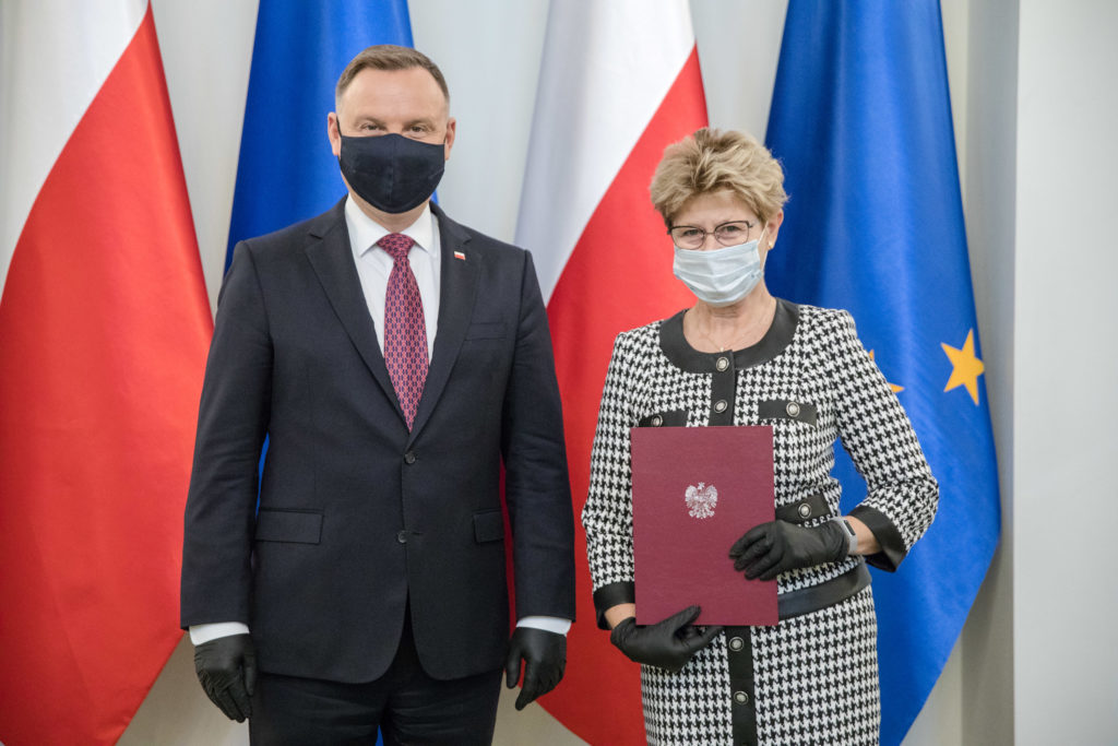Mężczyzna i kobieta w maseczkach stoją przed flagami Polski i UE. Kobieta trzyma dokument z godłem Polski.