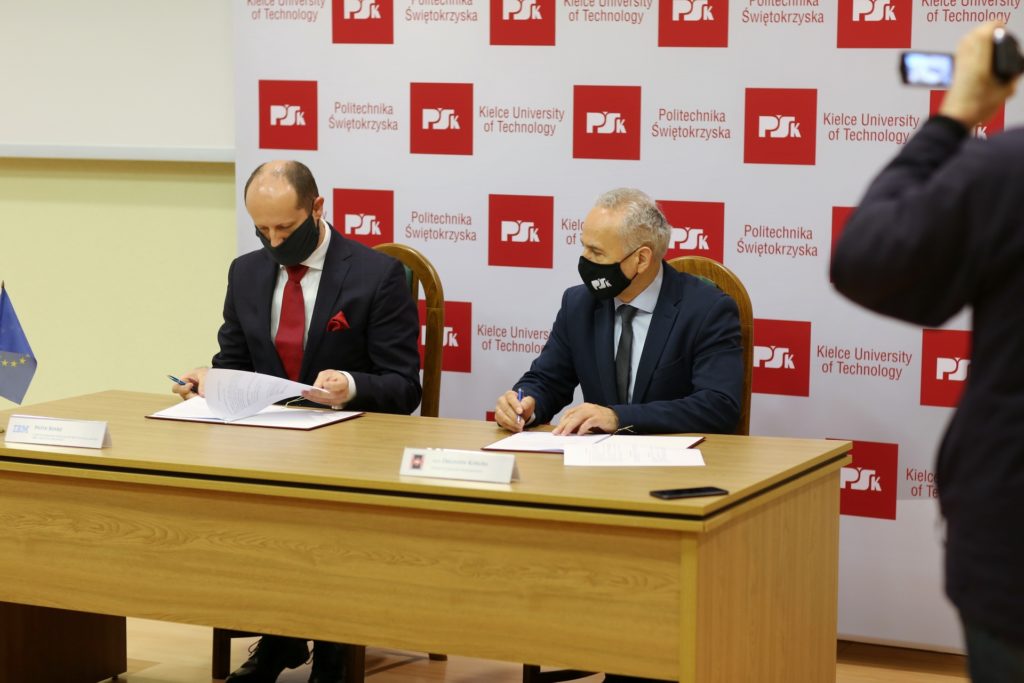 Dwóch mężczyzn w garniturach i maseczkach podpisuje dokumenty przy drewnianym stole. W tle baner Politechniki Świętokrzyskiej.