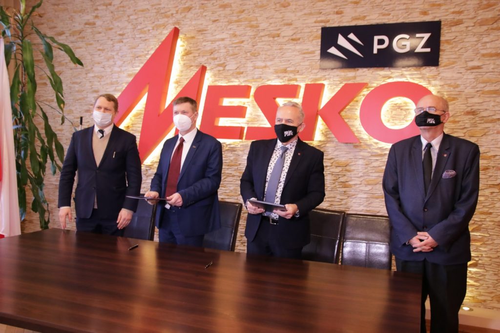 Czterech mężczyzn w garniturach i maseczkach stoi przed ścianą z logo MESKO i PGZ.