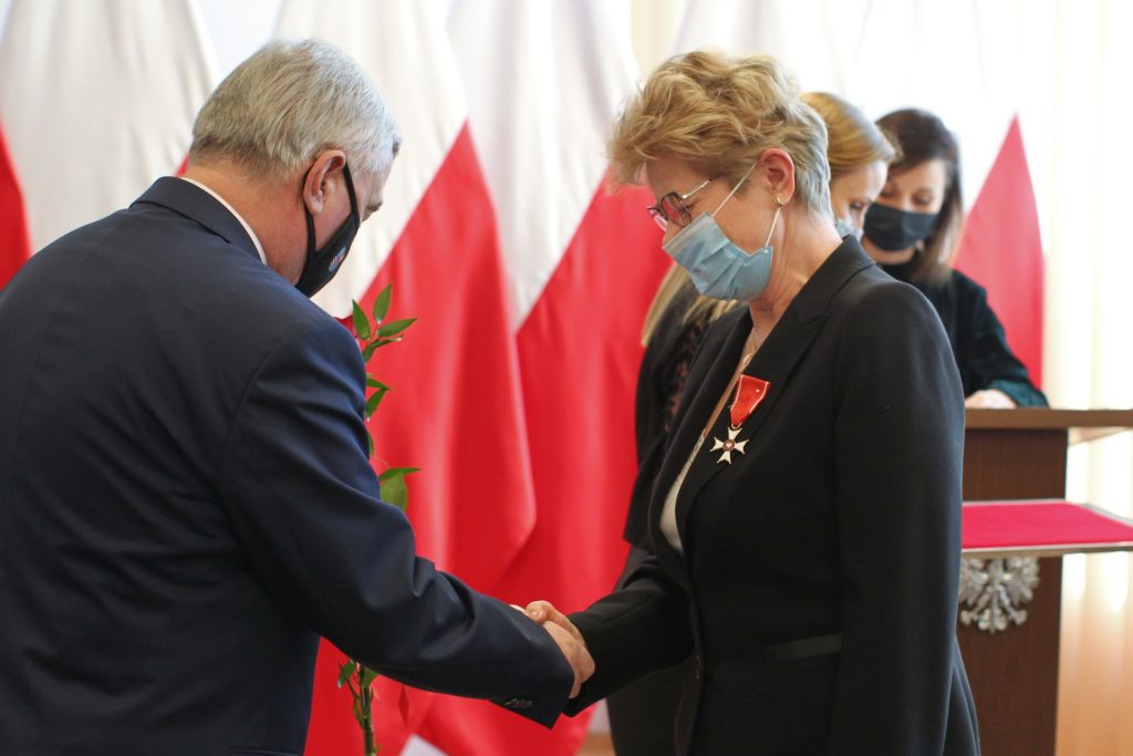Mężczyzna w garniturze podaje gałązkę kobiecie w czarnej marynarce z orderem. W tle polskie flagi.