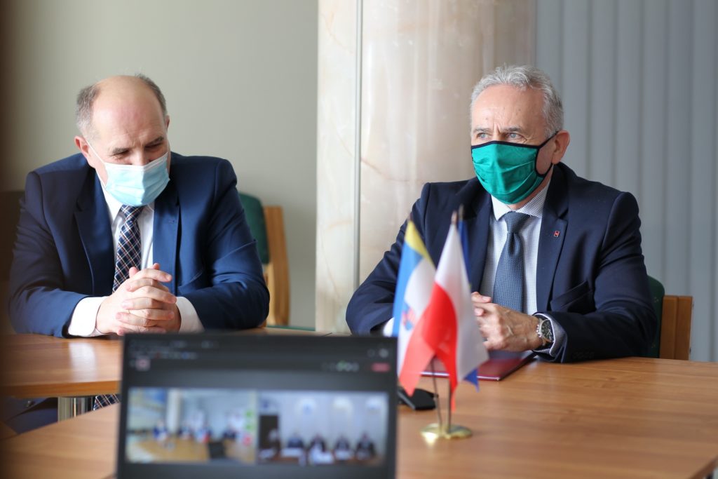 Dwóch mężczyzn w garniturach i maseczkach siedzi przy stole. Na stole flagi i laptop z wideokonferencją.