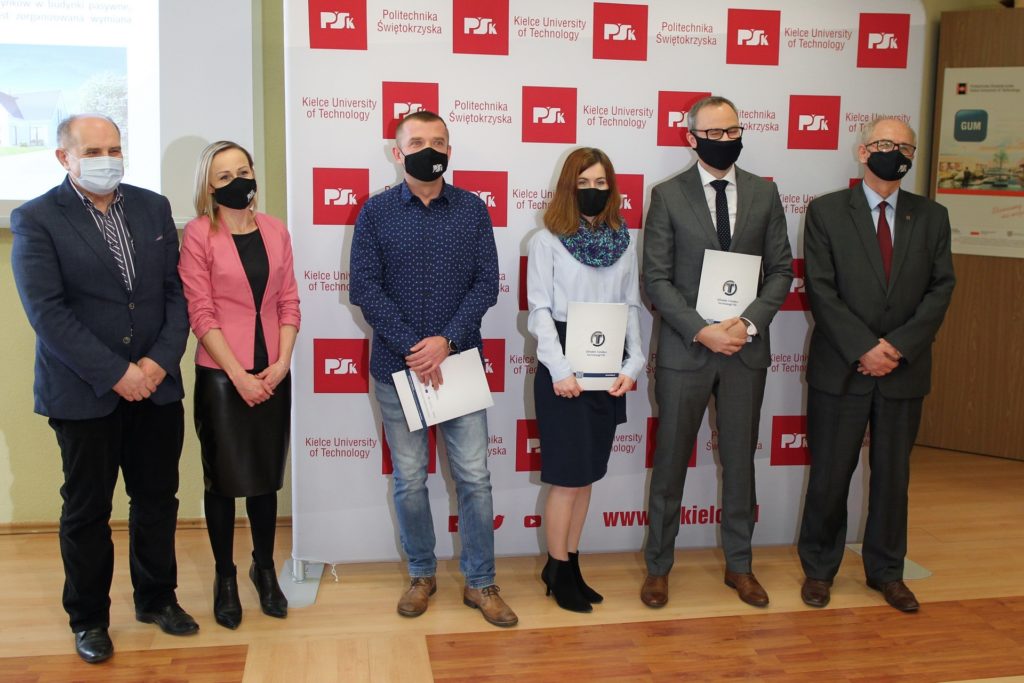 Osoby w maseczkach stoją przed banerem Politechniki Świętokrzyskiej. Kluczowe obiekty: ludzie, baner, dokumenty.