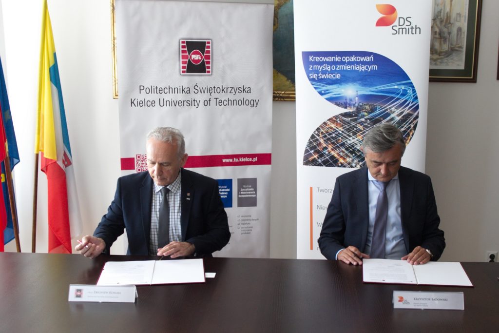 Dwóch mężczyzn w garniturach podpisuje dokumenty przy stole. W tle banery Politechniki Świętokrzyskiej i DS Smith.