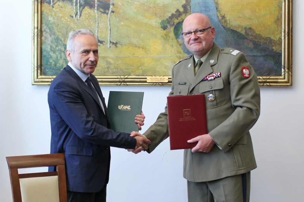 Dwóch mężczyzn, jeden w garniturze, drugi w mundurze wojskowym, wymienia uścisk dłoni. Trzymają dokumenty.